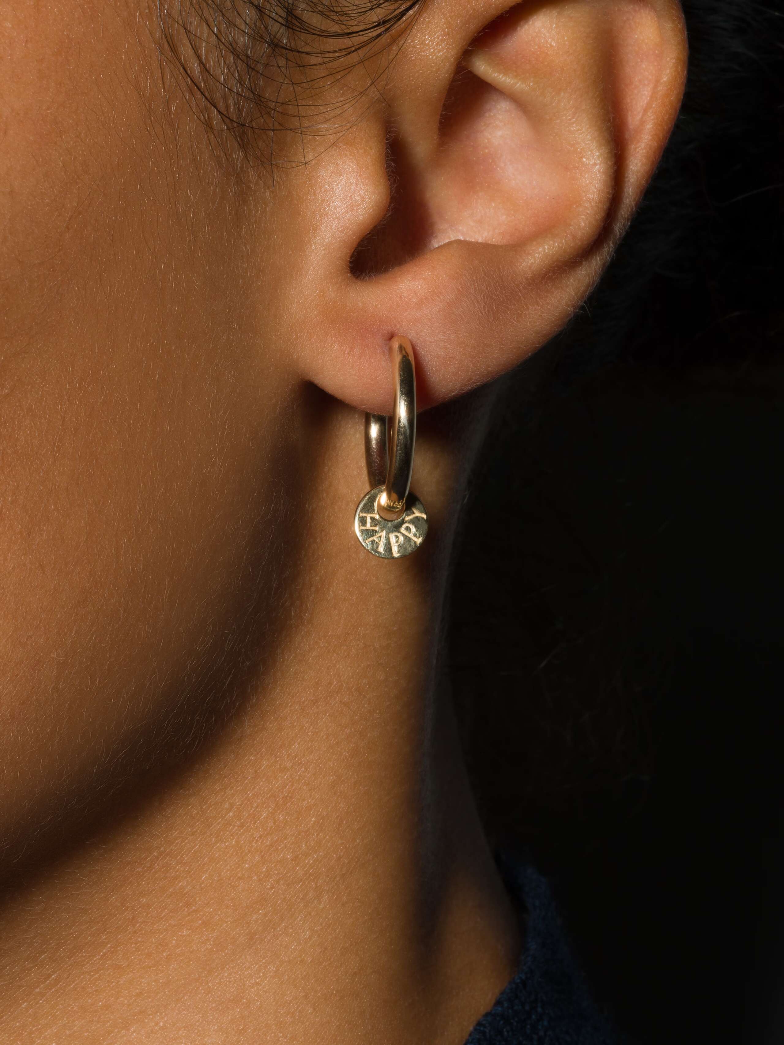 model wears earring in creole shape with engraved ("happy") small round tag in gold 