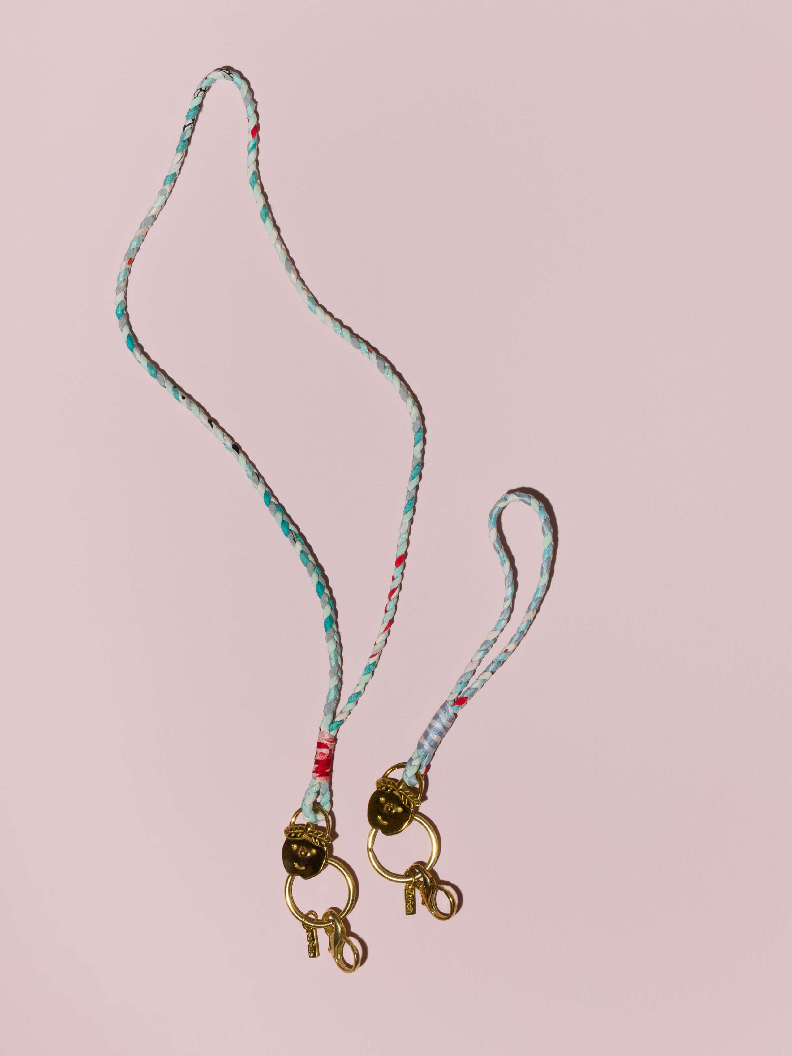 two braided keychains in light blue shades in silk with a brass tag in shape of a cheeky boys' face