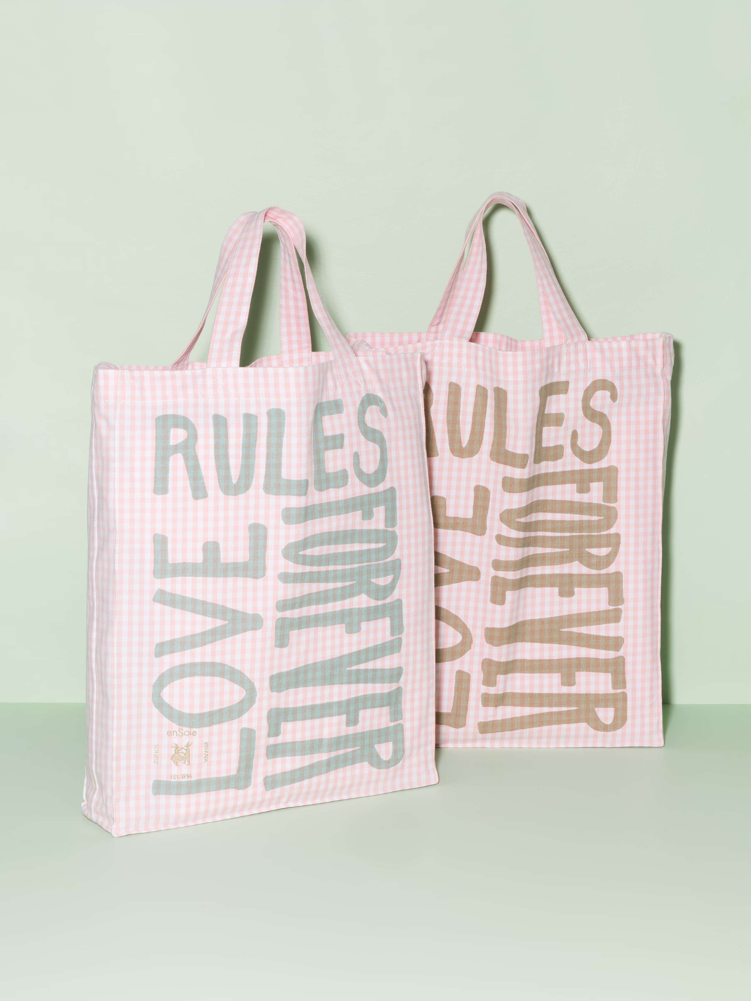 Two cotton tote bags with a pink vichy pattern and a mint and beige print that says "Love rules forever"