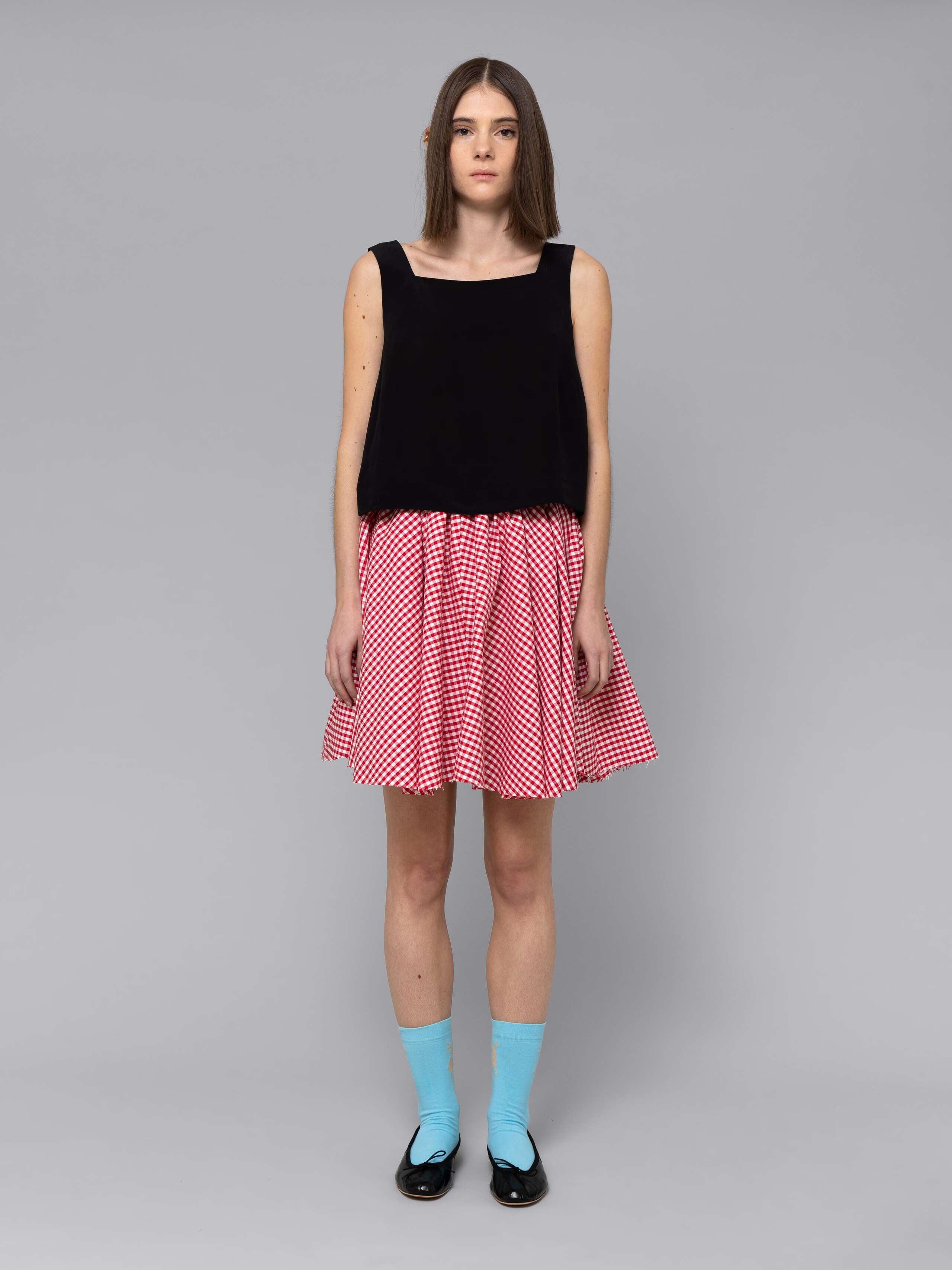 Female model is wearing a combination of a boxy black silk top and a cute short cotton skirt  featuring a beautiful red vichy check pattern