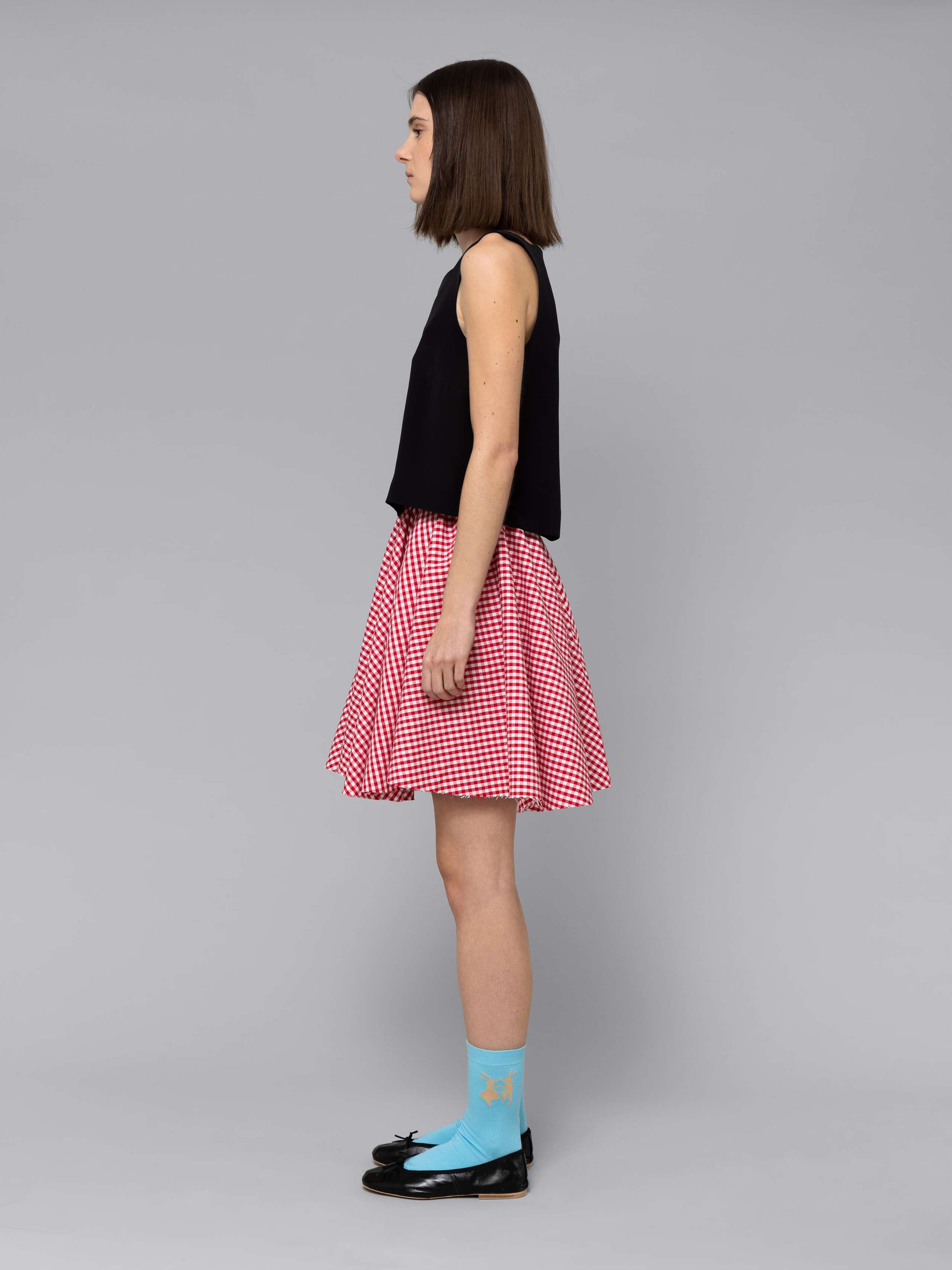 Female model is wearing a combination of a boxy black silk top and a cute short cotton skirt  featuring a beautiful red vichy check pattern