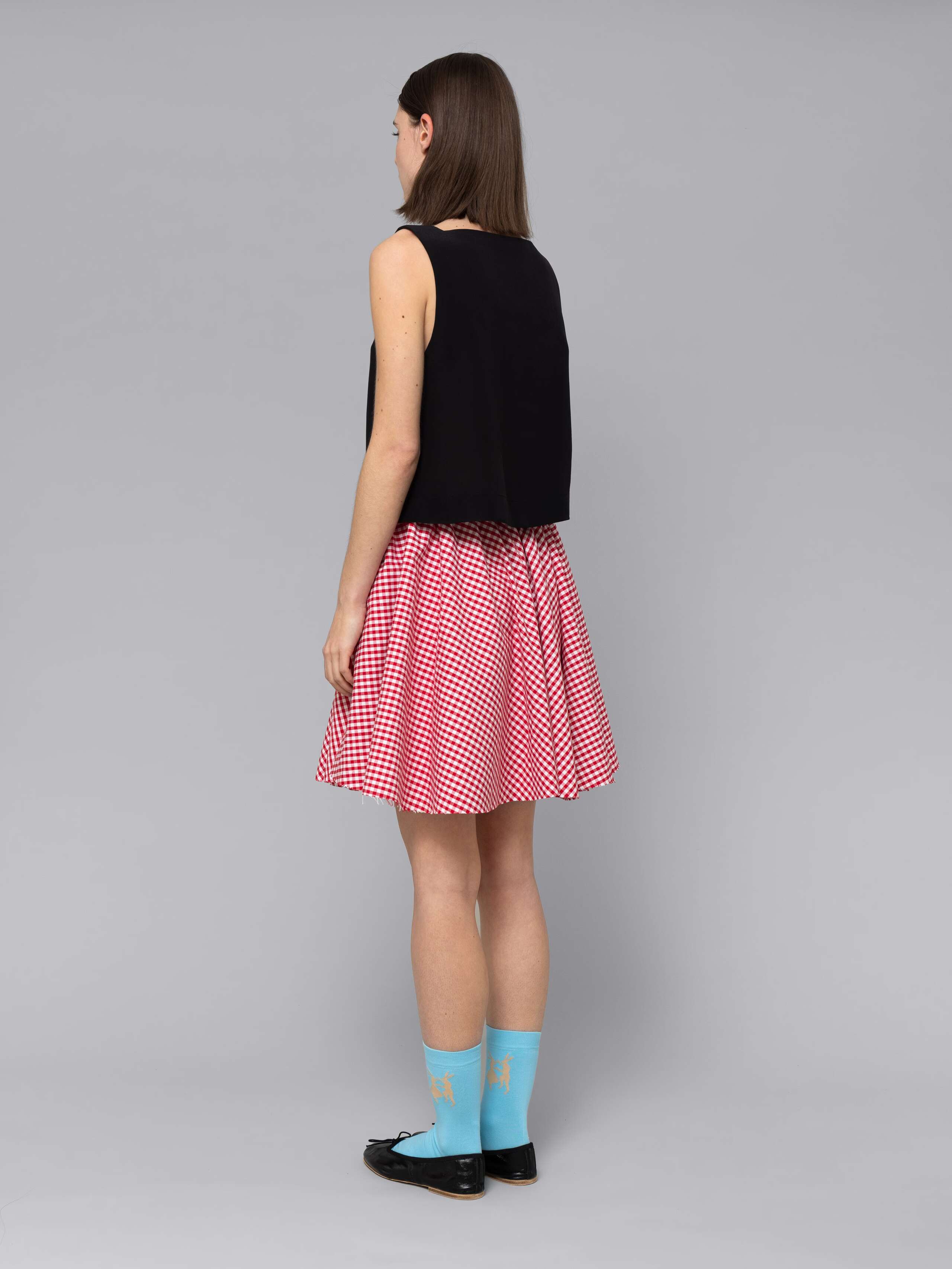 Female model is wearing a combination of a boxy black silk top and a cute short cotton skirt  featuring a beautiful red vichy check pattern
