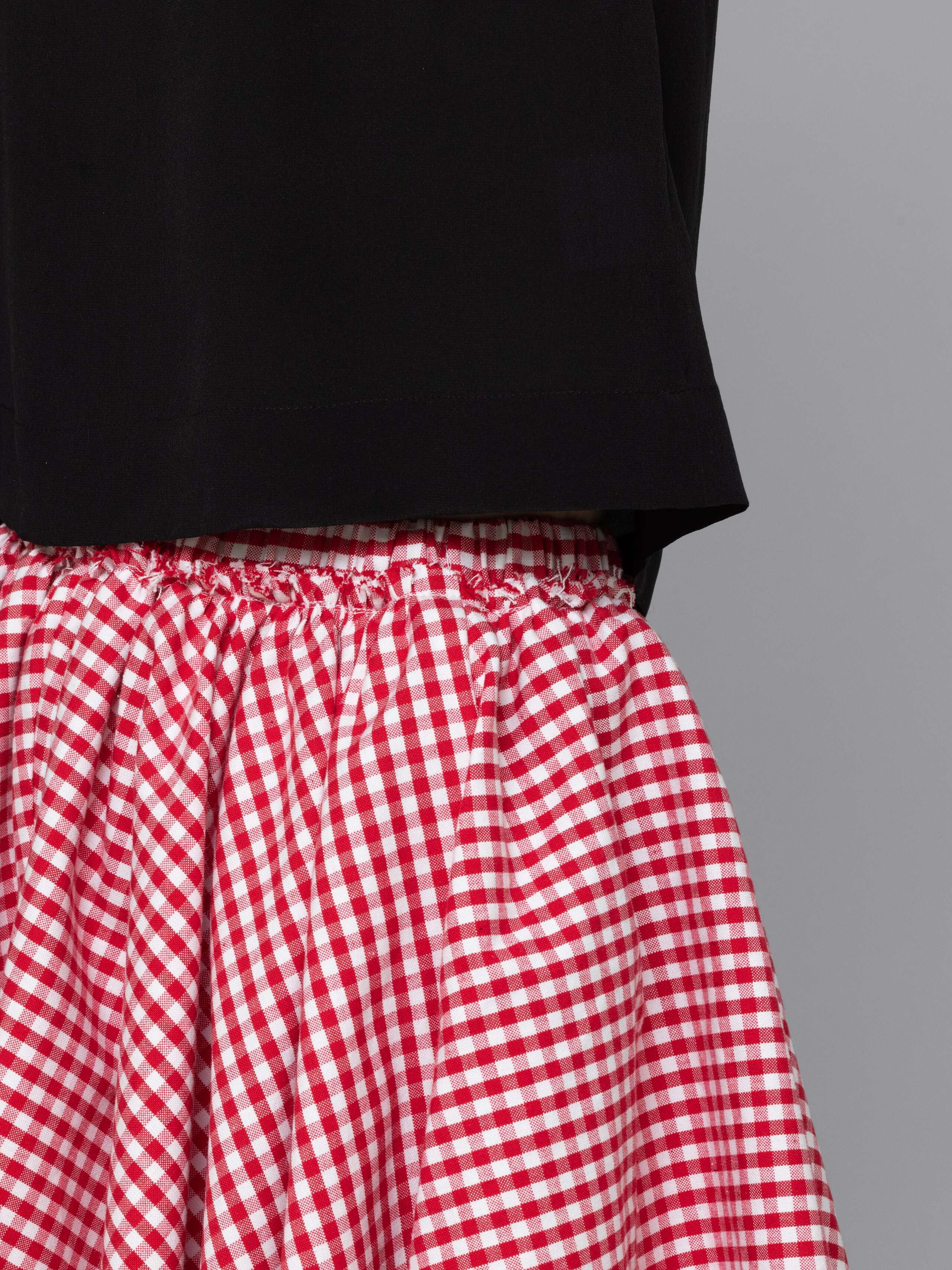 Female model is wearing a combination of a boxy black silk top and a cute short cotton skirt featuring a beautiful red vichy check pattern