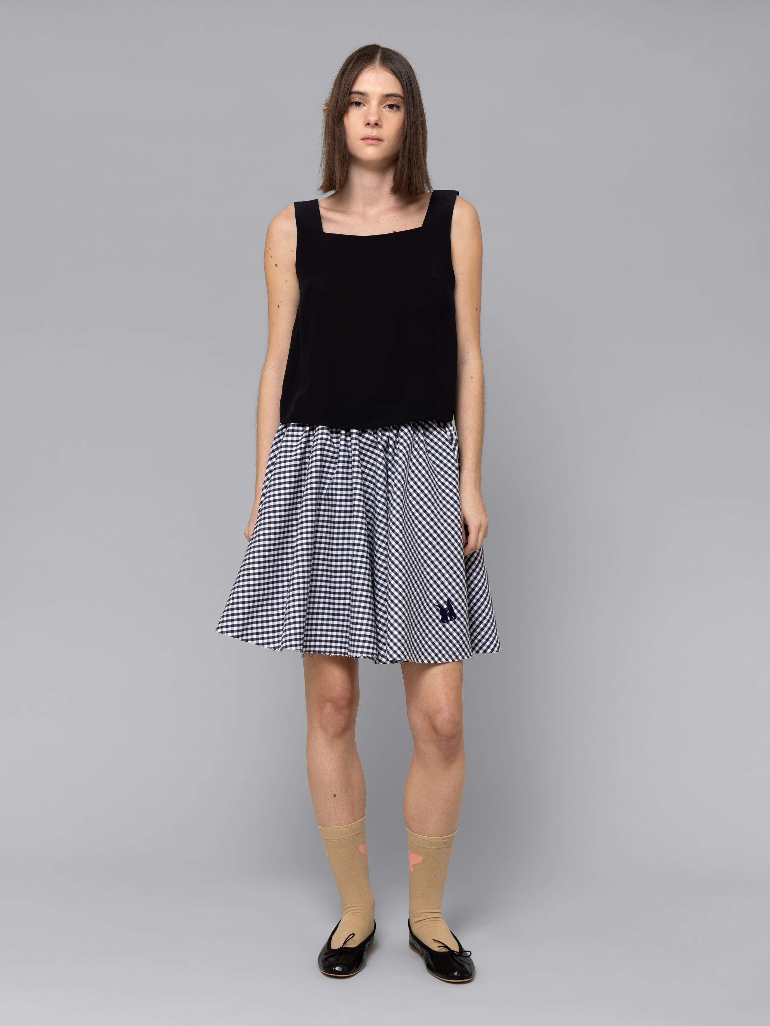 Female model is wearing a combination of a boxy black silk top and a cute short cotton skirt  featuring a beautiful navy vichy check pattern