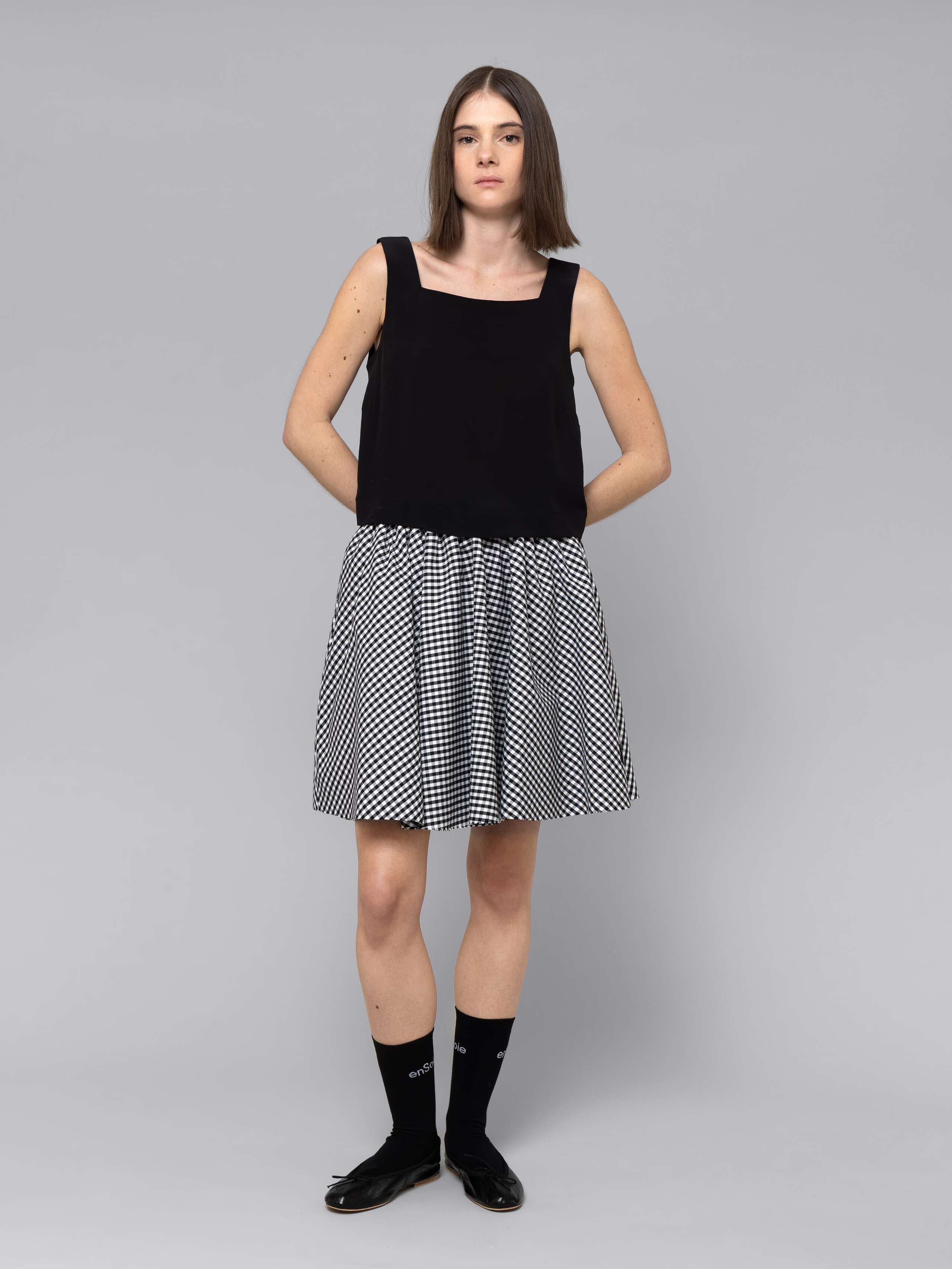 Female model is wearing a combination of a boxy black silk top and a cute short cotton skirt  featuring a beautiful black vichy check pattern