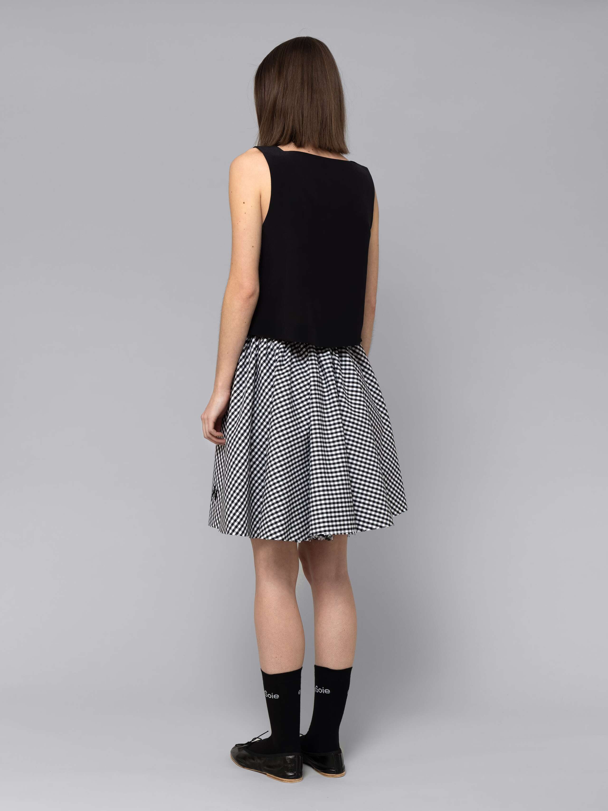 Female model is wearing a combination of a boxy black silk top and a cute short cotton skirt  featuring a beautiful black vichy check pattern
