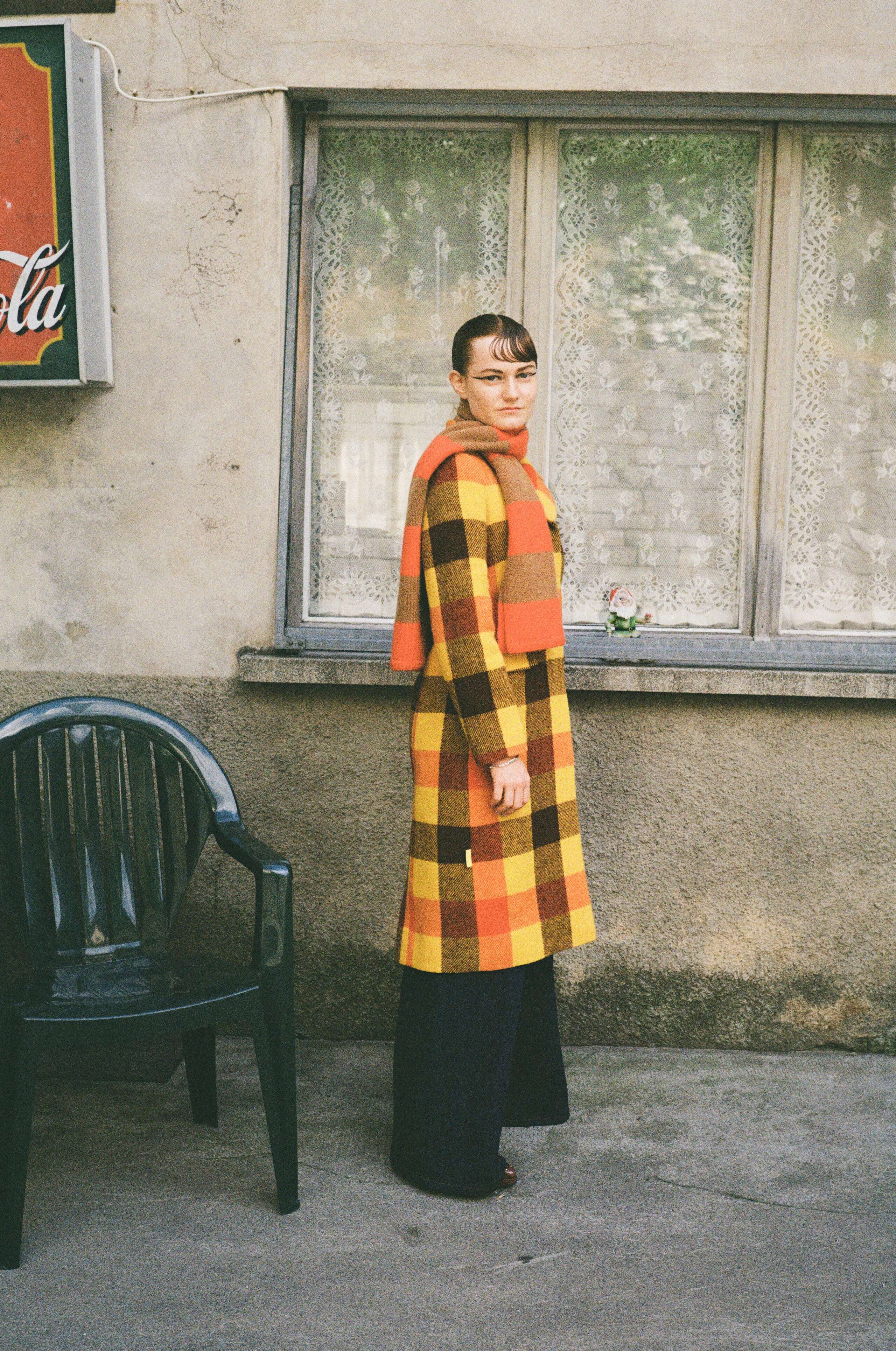 Picture of a female model wearing a colourful wool coat, standing in front of a restaurant